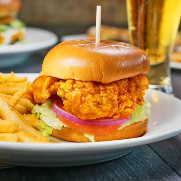 Crispy chicken sandwich with lettuce, tomato, onion, and fries, with a beer in the background.