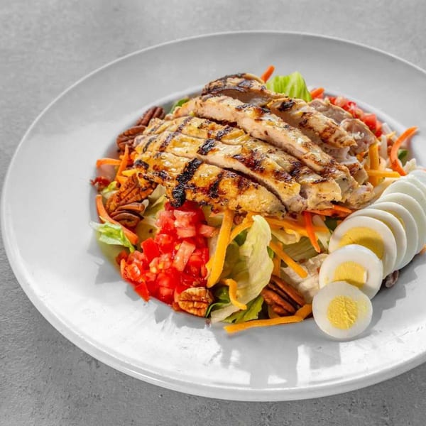 Grilled chicken salad with lettuce, tomatoes, carrots, pecans, and sliced hard-boiled eggs on a white plate.