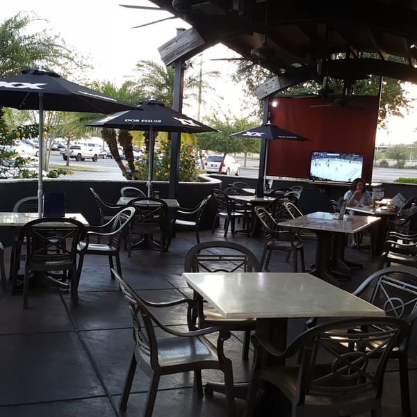Outdoor patio at Tampa Joe's with tables, chairs, umbrellas, and a TV showing a sports game.