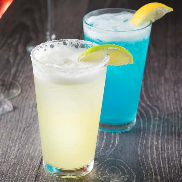 Two vibrant cocktails, a yellow margarita with a lime and a blue drink with a lemon, on a dark wooden table.