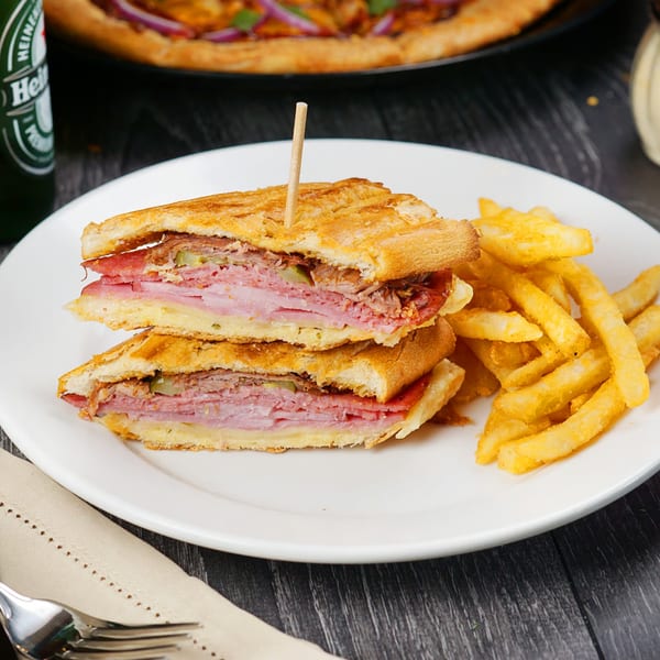 A Cuban sandwich with fries on a white plate, with a beer bottle and pizza in the background.